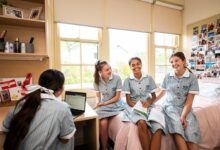 Four teenaged girls in blue and white school uniforms sitting in a school boarding house bedroom, laughing