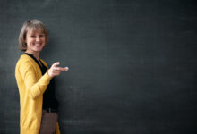 Young teacher with piece of chalk is standing near blackboard in a classroom.