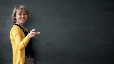 Young teacher with piece of chalk is standing near blackboard in a classroom.